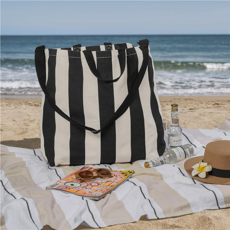 Black and white striped tote bag on a beach with a magazine, sunglasses, and a hat.