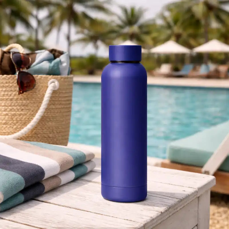 Blue water bottle on a table by a poolside with palm trees and lounge chairs in the background.