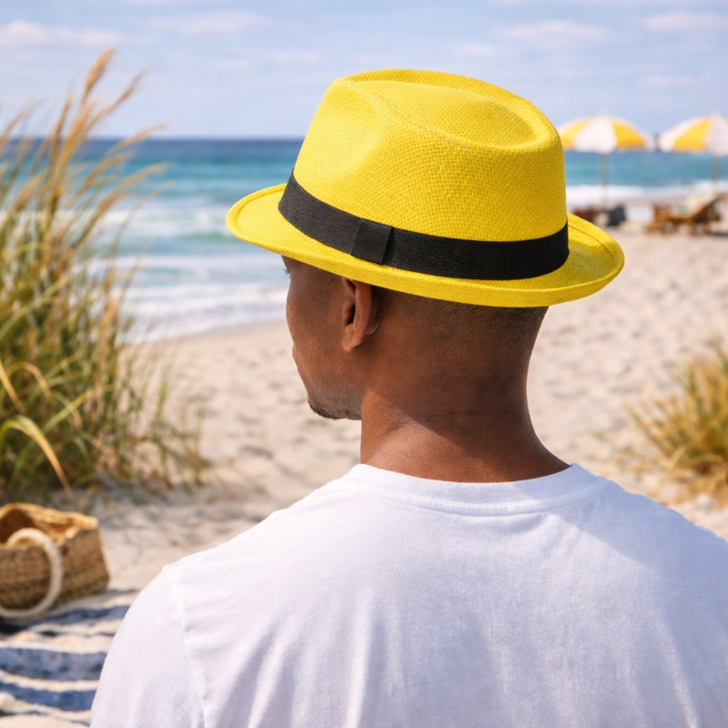 Person wearing a yellow fedora hat with black band on a beach with ocean view