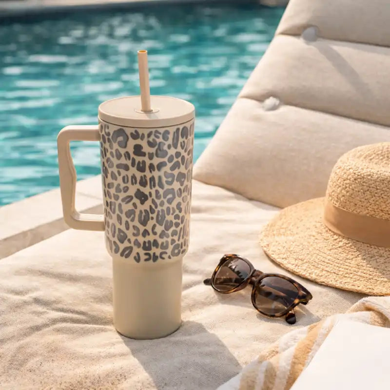 Leopard print mug with straw, sunglasses, and hat by a poolside.