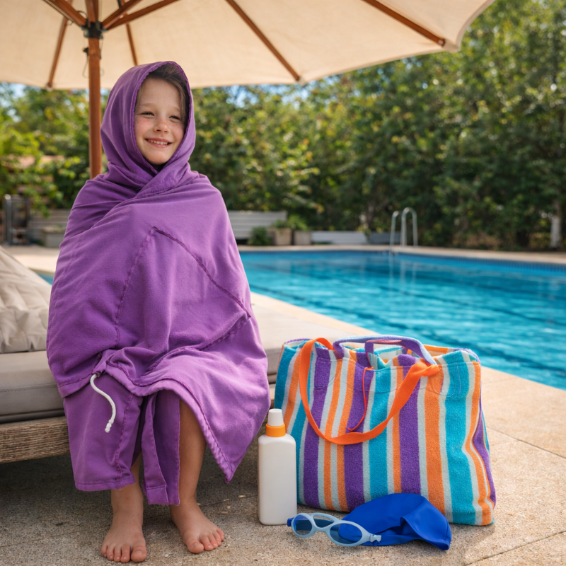 Child wrapped in a purple TowelTote by a poolside with a striped bag and sunglasses.